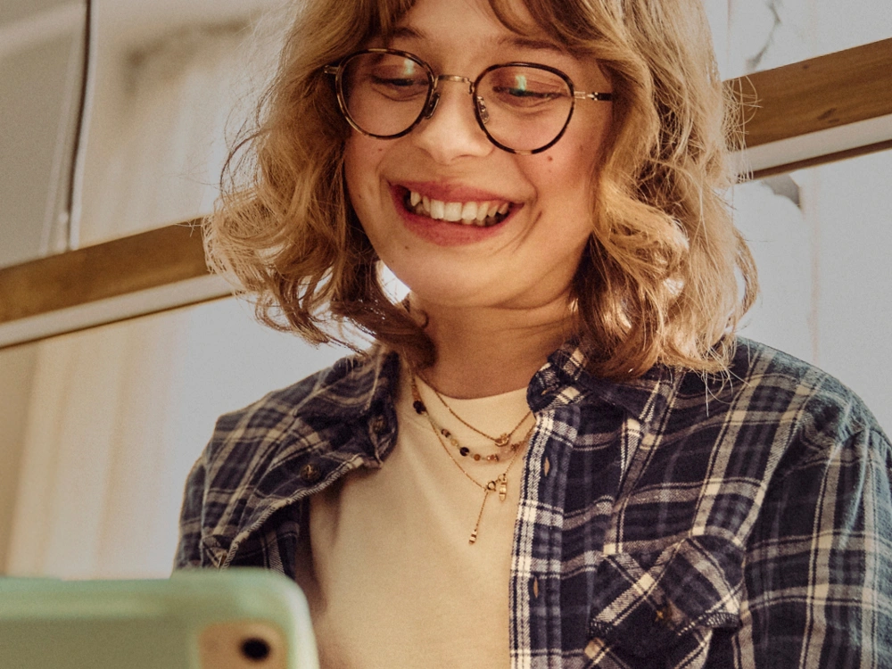 Smilende person med briller bruger mobiltelefon i hyggelige og lyse omgivelser.